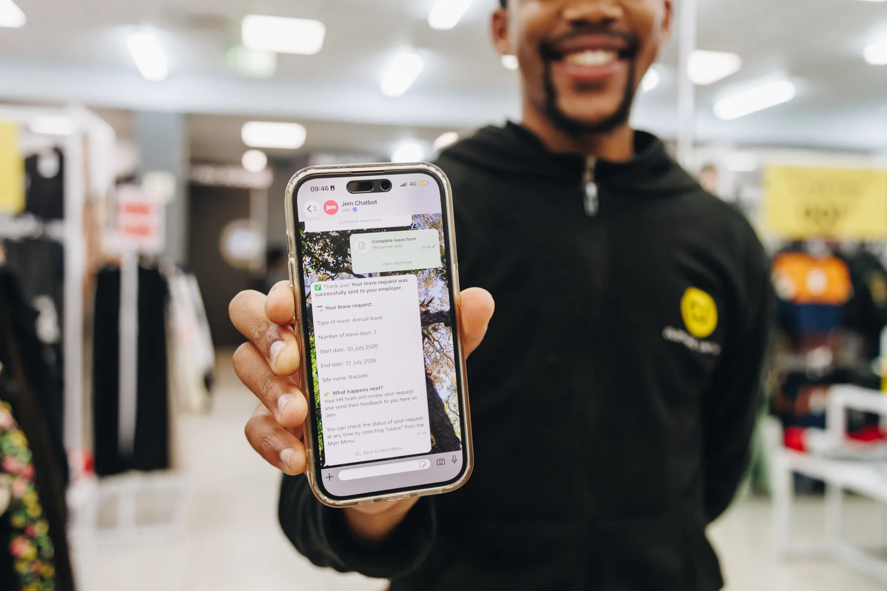A retail employee in a branded hoodie holding up a phone showing the Jem chatbot’s confirmation that their leave request has been sent to their employer.