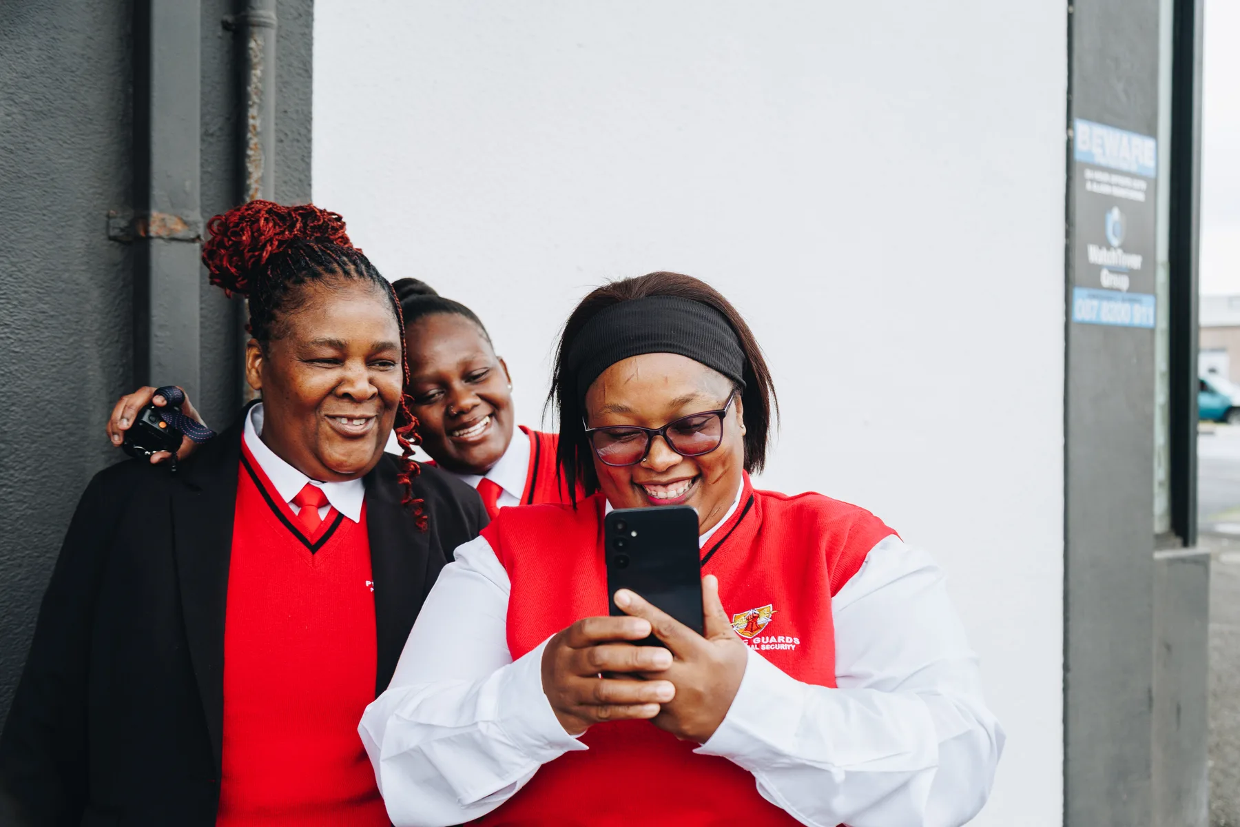 Three security guards in red and white uniform standing outside their site, laughing at a message on one of their phones.