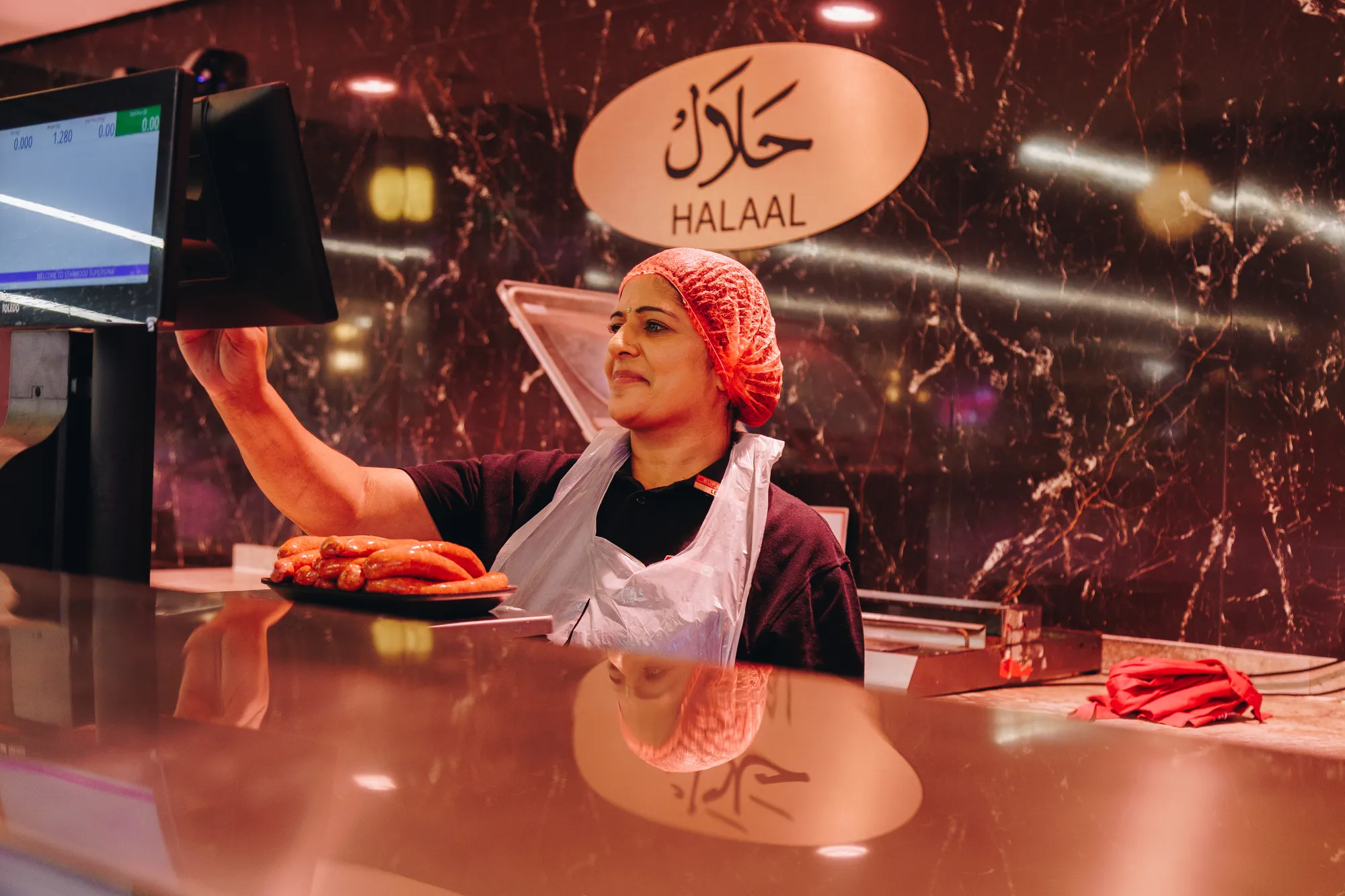 A Superspar butchery employee in a red hairnet and white apron reaches up to place a tray of meat on the counter beneath a halaal sign