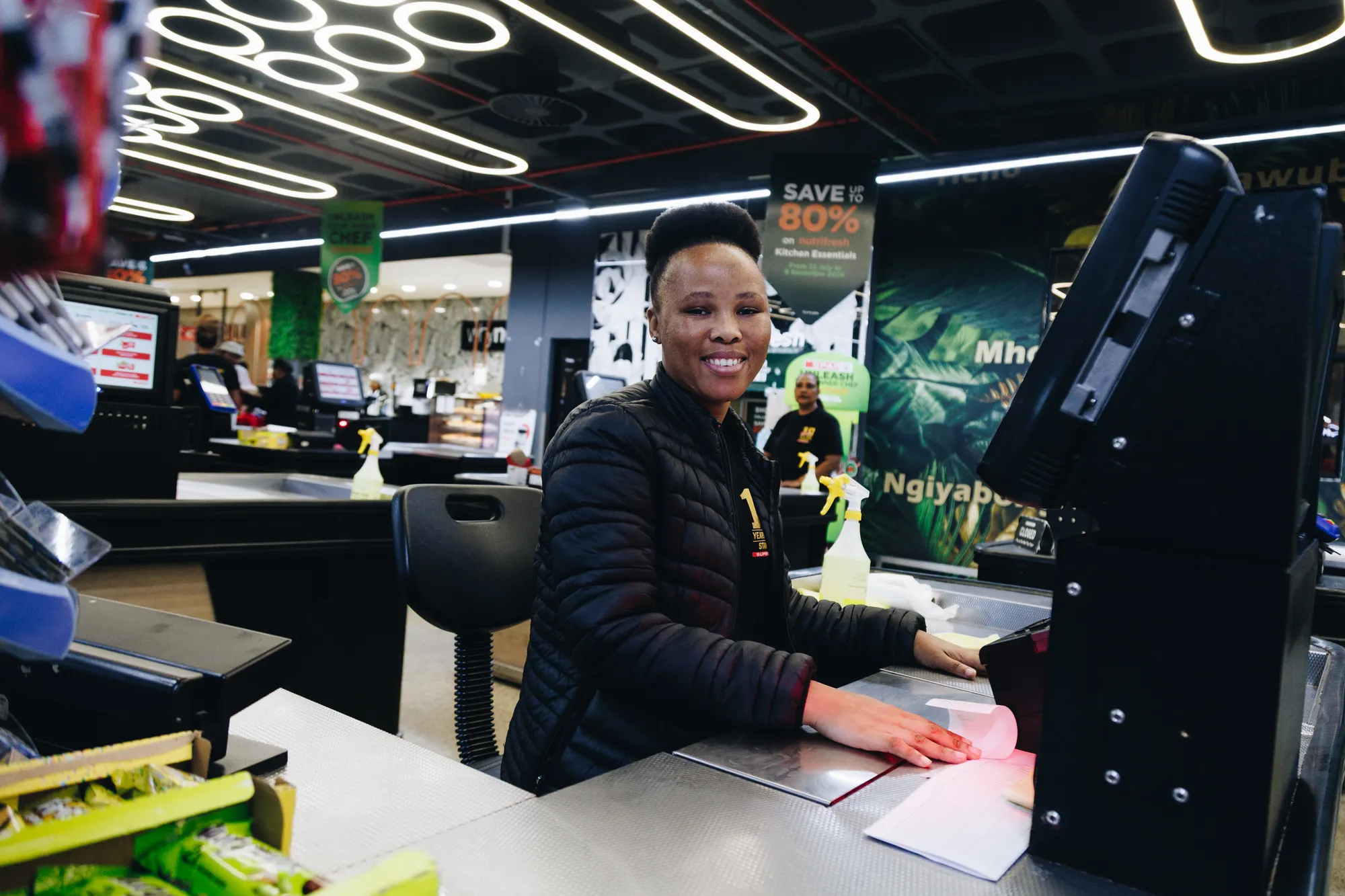 A smiling Superspar cashier in a black jacket sits at her till, one hand resting on the counter