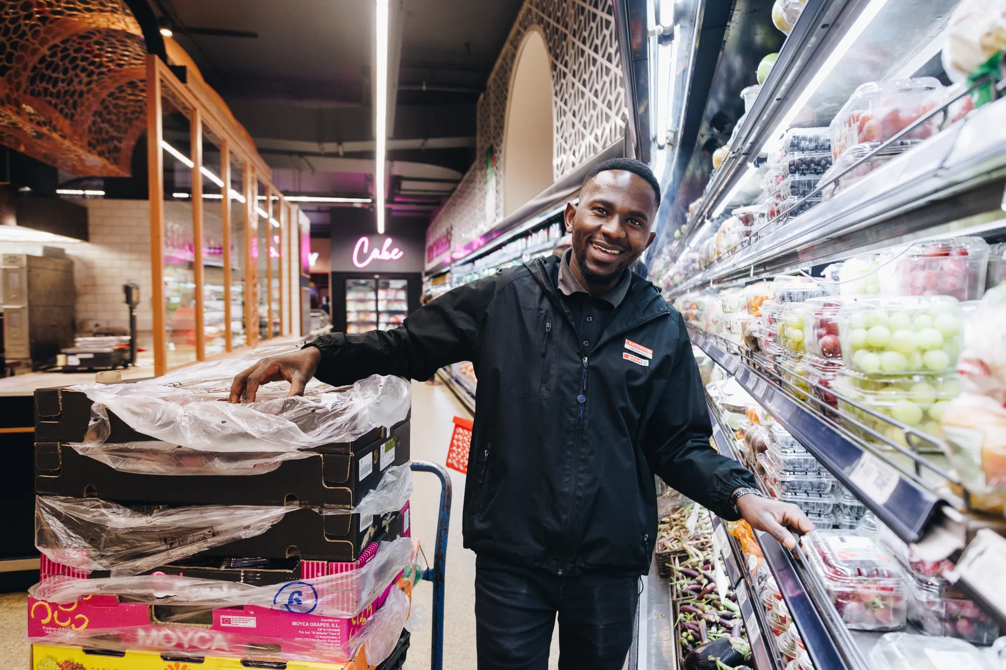 A smiling Superspar employee in a black jacket reaches into the fresh produce display in a bright, well-stocked store