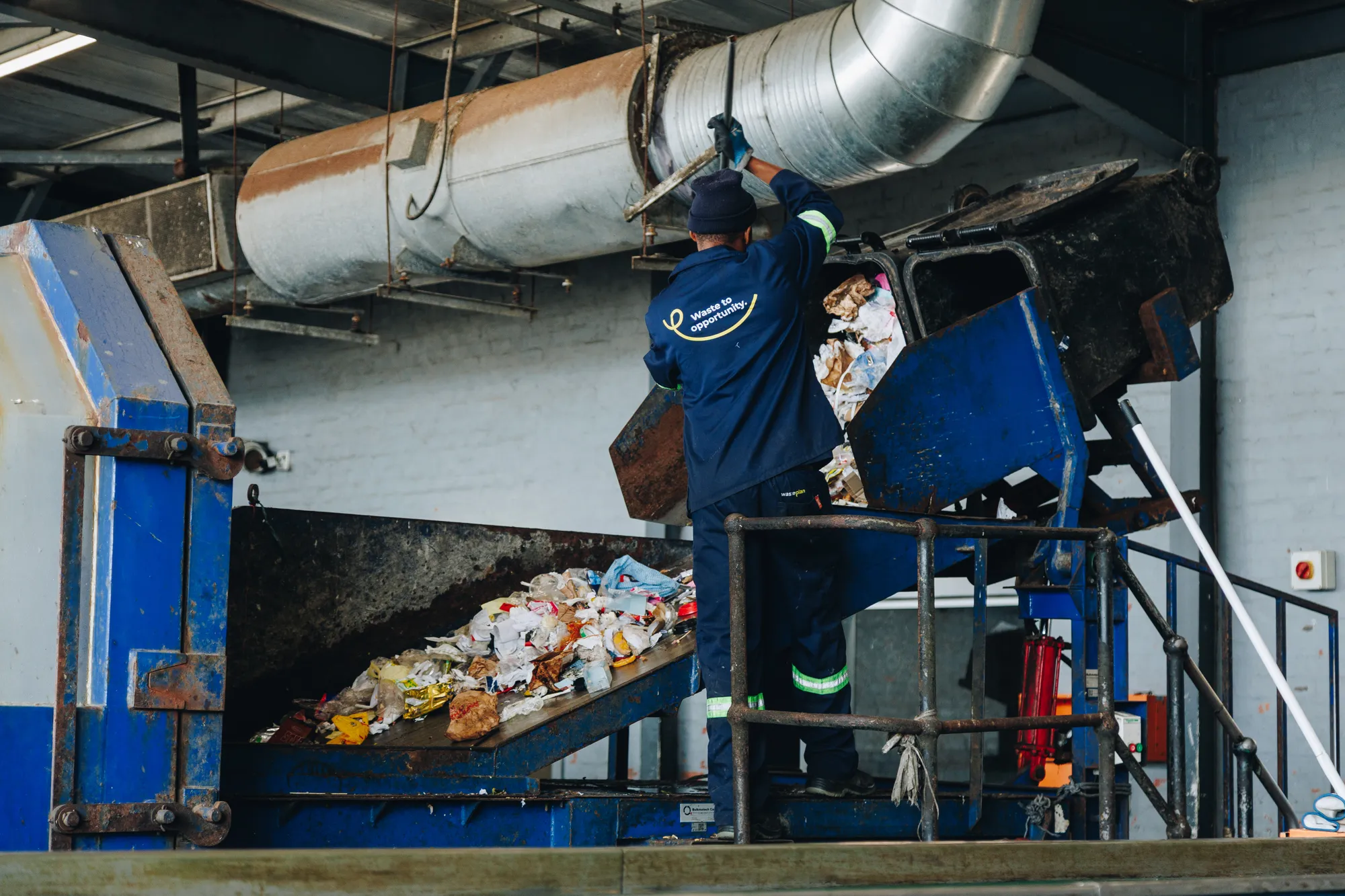 A WastePlan worker in navy overalls with high-visibility bands stands at a blue chute, feeding sorted material into a machine