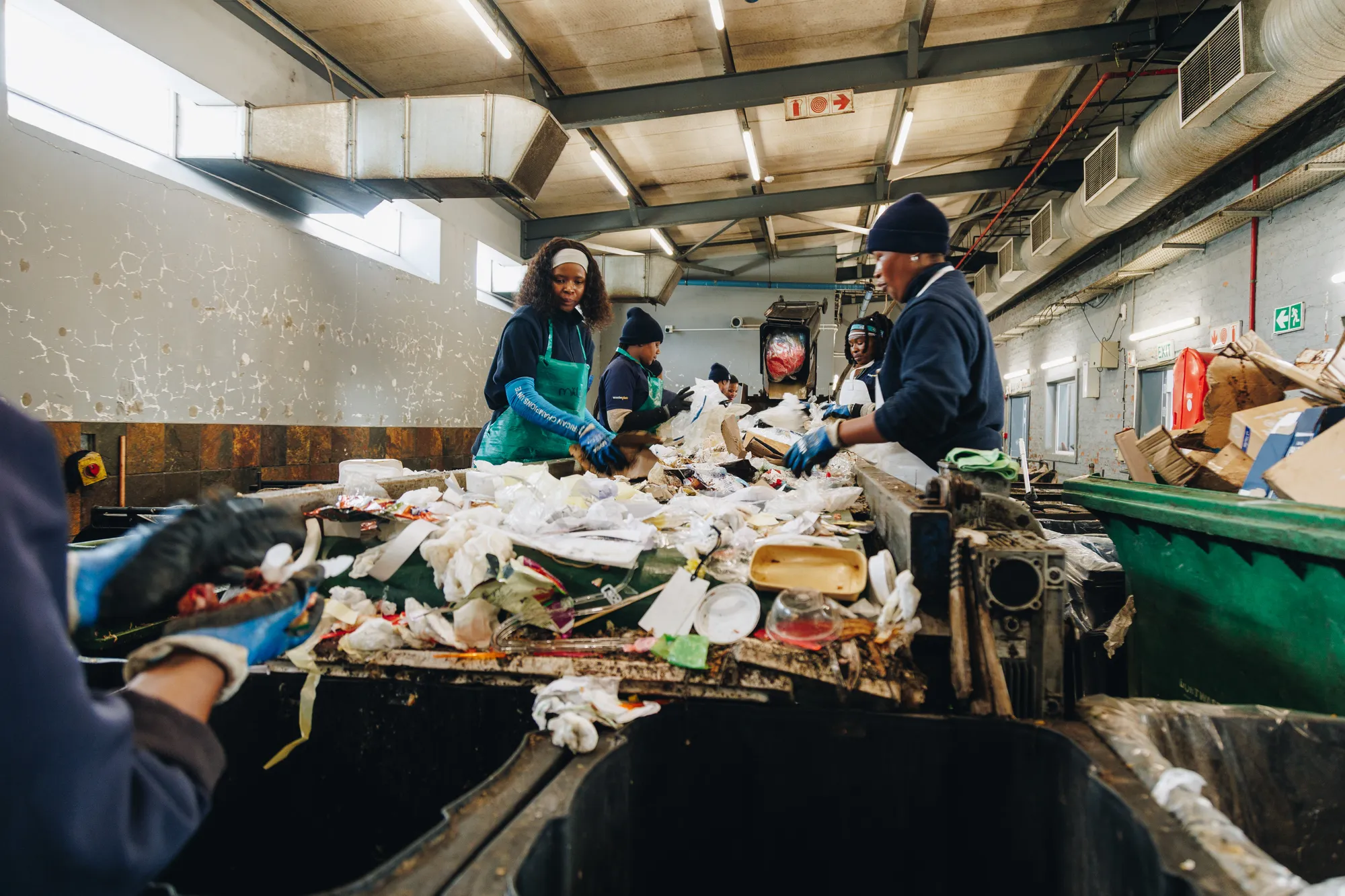 A crew of WastePlan sorters working along a conveyor belt inside a sorting hall, in navy fleeces, green aprons and blue gloves