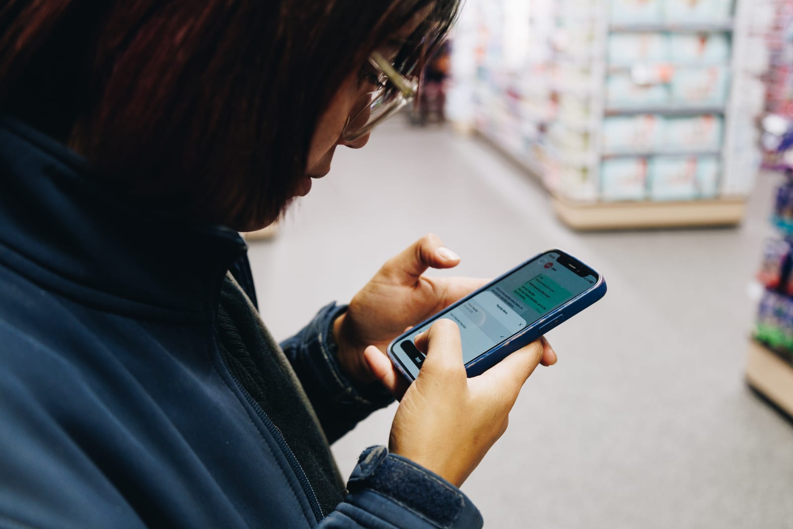 Over-the-shoulder view of a shop worker in a navy fleece reading a WhatsApp chat on her phone in a store aisle