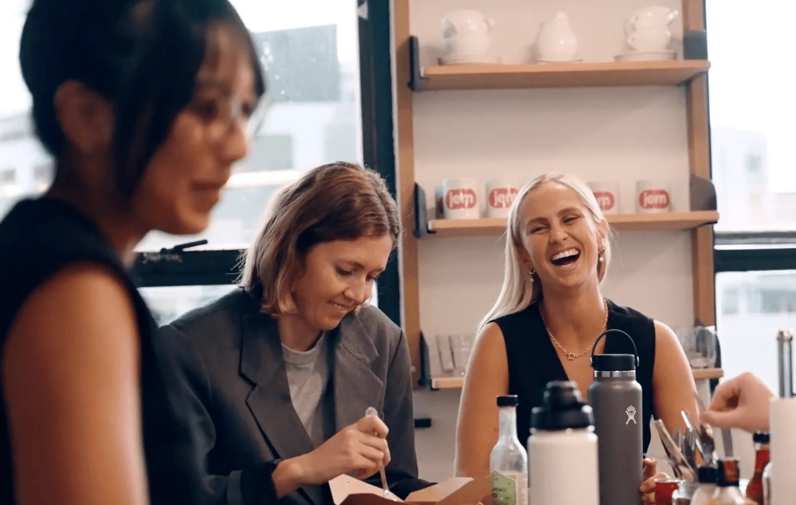Three Jem colleagues laughing together over lunch in the office kitchen, Jem mugs on the shelf behind them