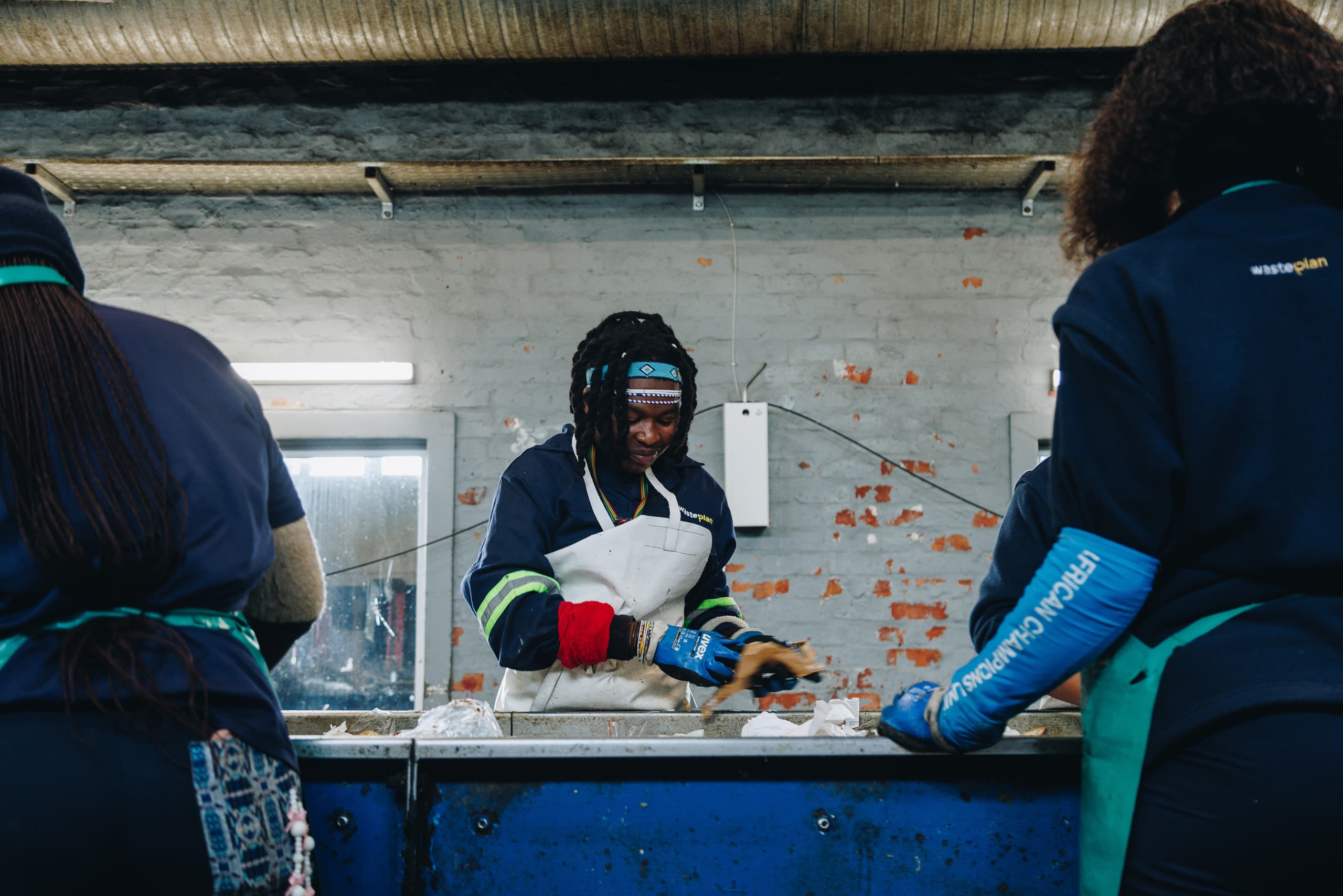 A WastePlan sorter in a white apron and blue gloves smiling as she picks through material at a sorting belt, framed by two colleagues in navy WastePlan fleeces in the foreground