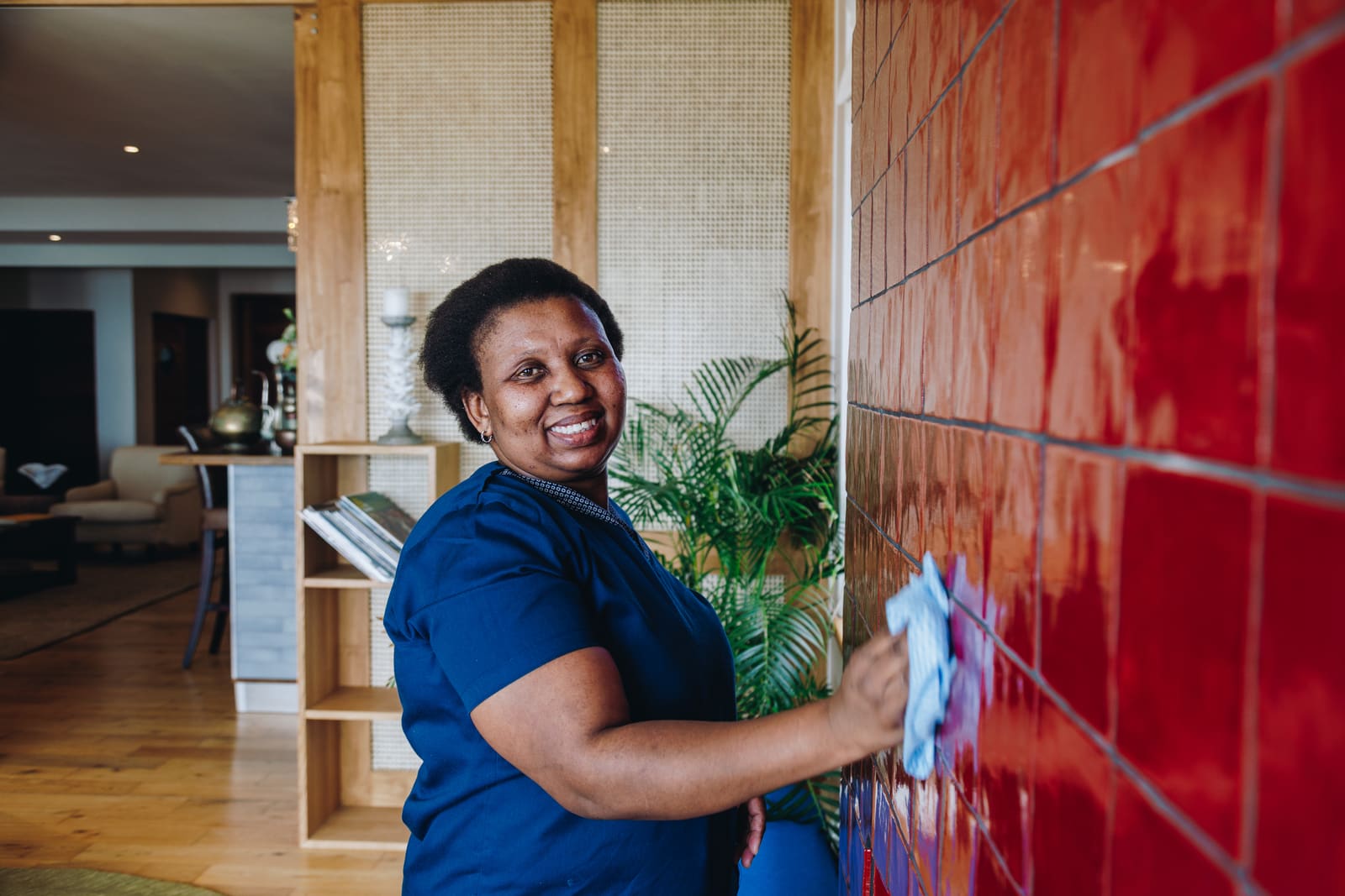 A smiling cleaner in a navy uniform wiping a glossy red tiled wall with a blue cloth in a hotel lounge