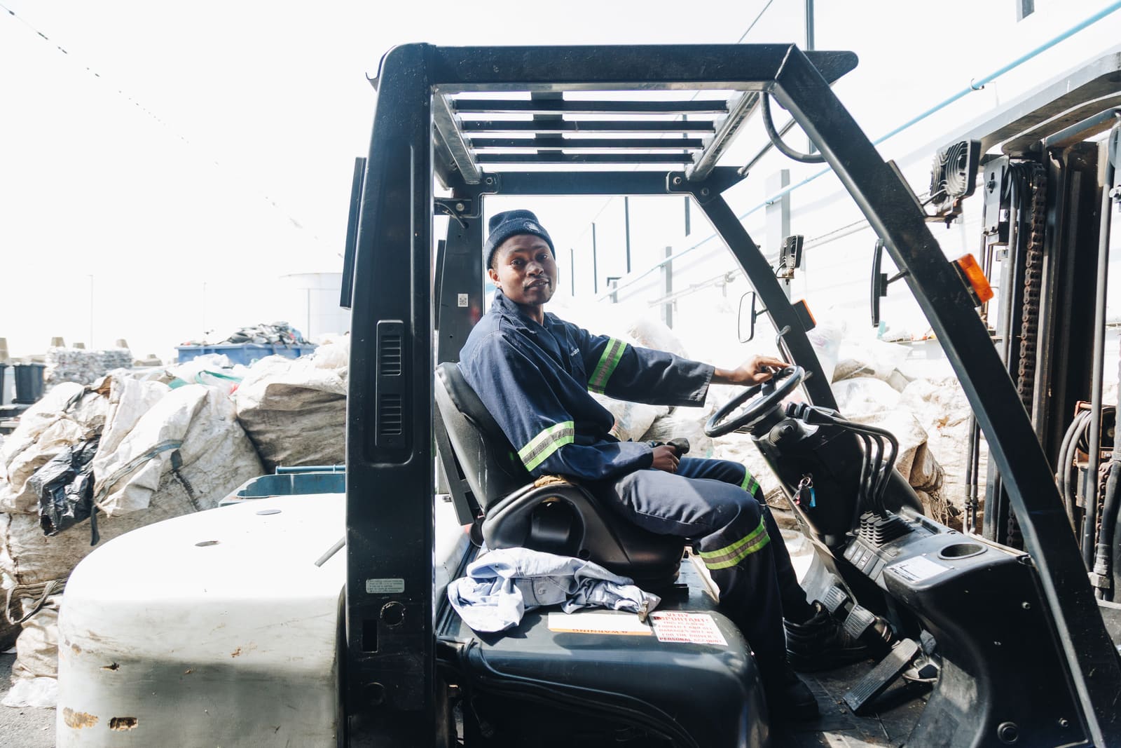 A worker in a navy uniform and beanie seated at the wheel of a forklift beside stacked bales of material in a yard
