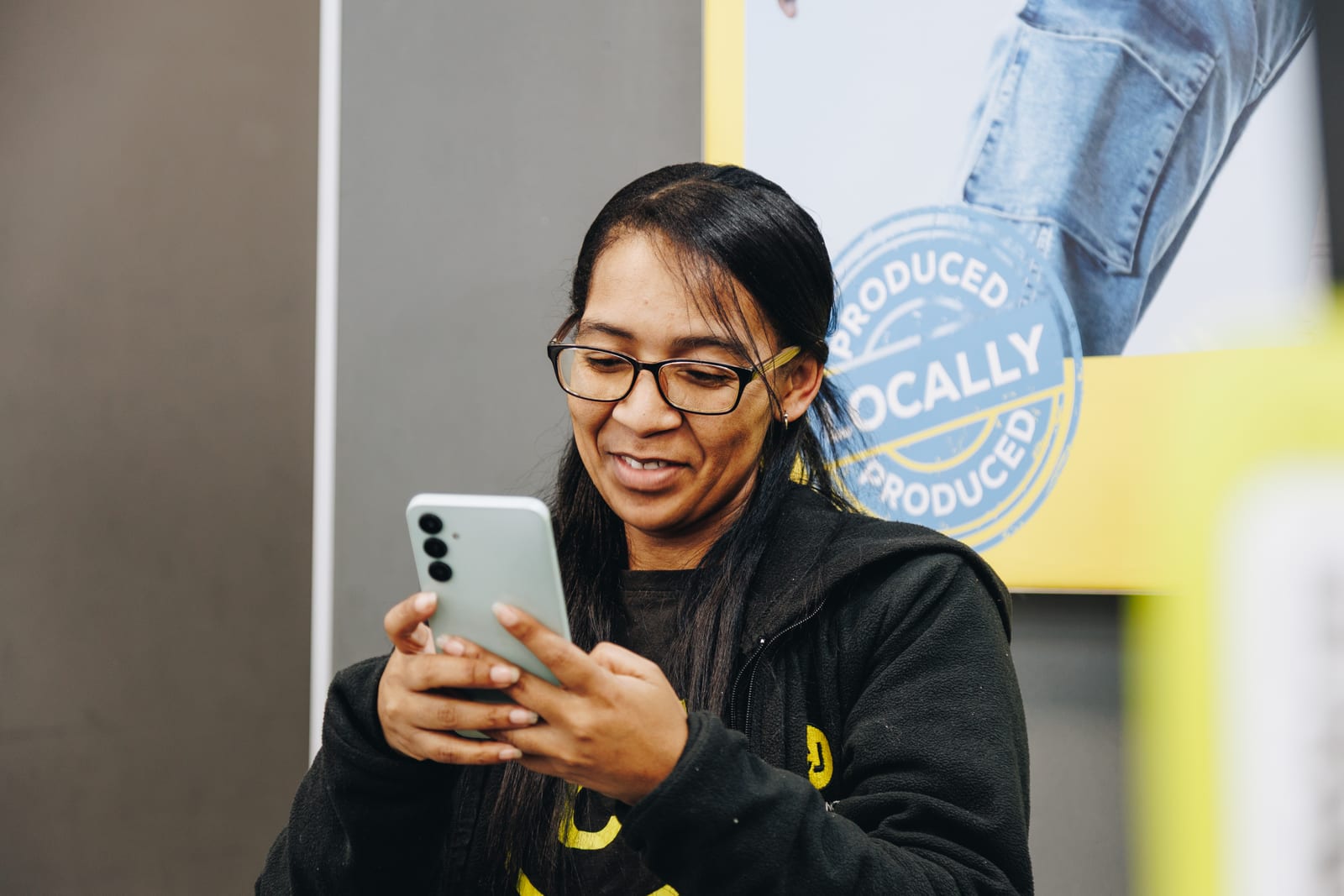 A retail worker in a branded fleece smiling at her phone, a shop poster on the wall behind her