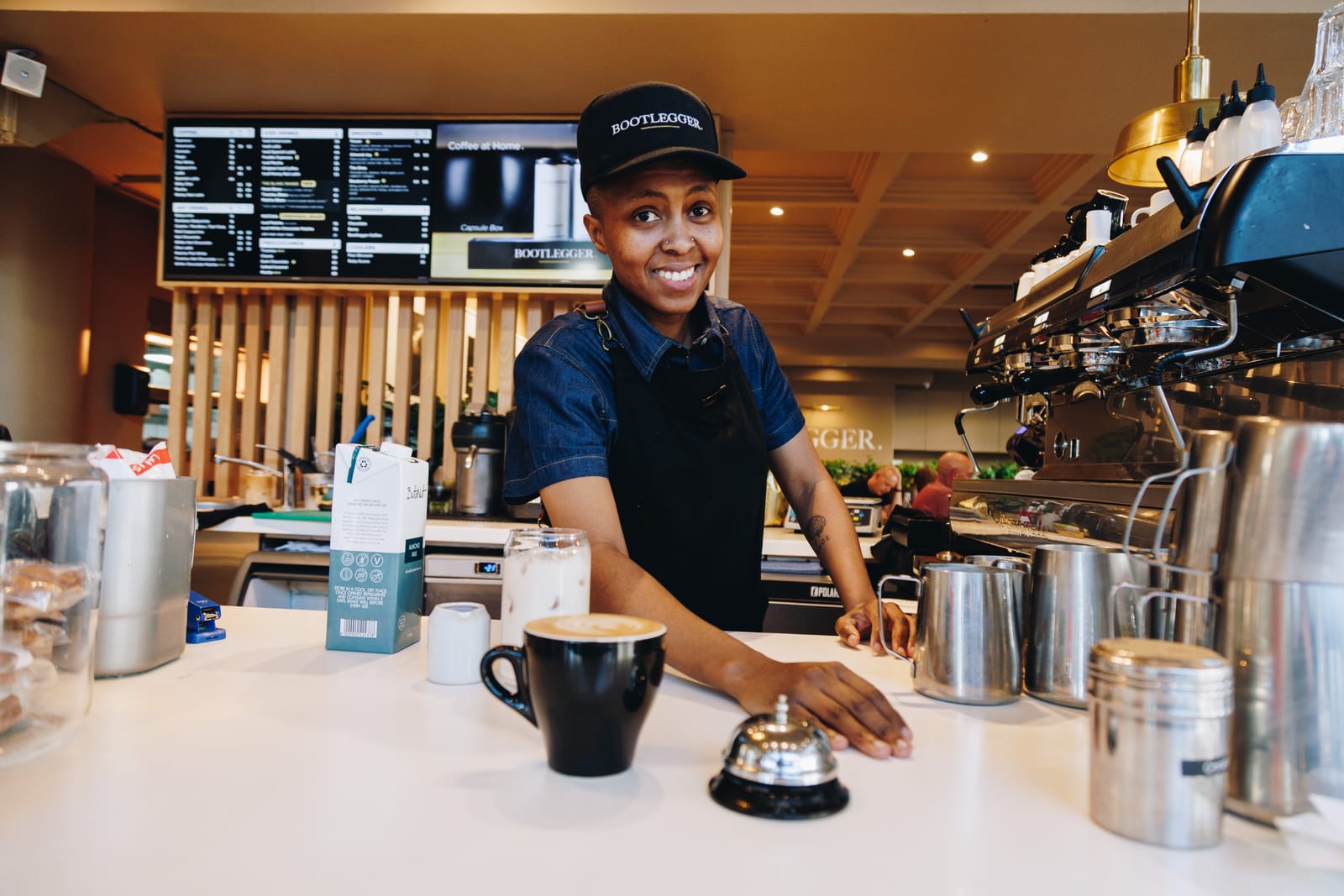 A barista in a cap and apron leaning on the counter beside a coffee and a service bell