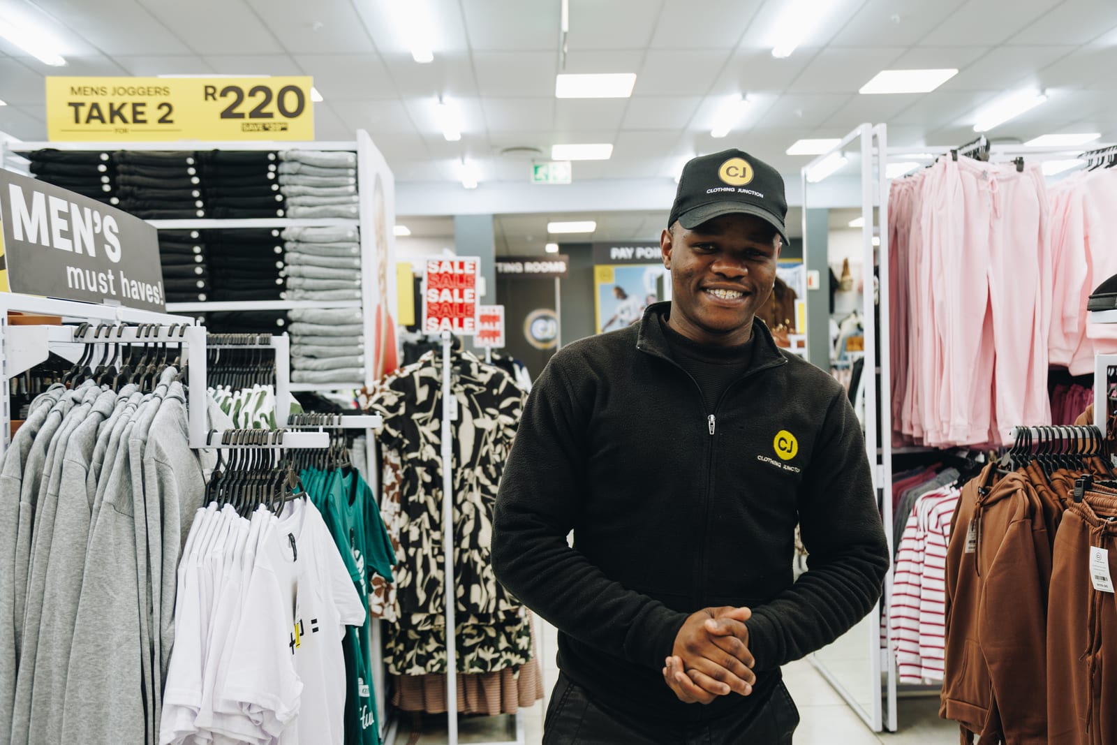 A shop assistant in a branded cap and fleece smiling on the floor of a clothing store, sale signs behind him