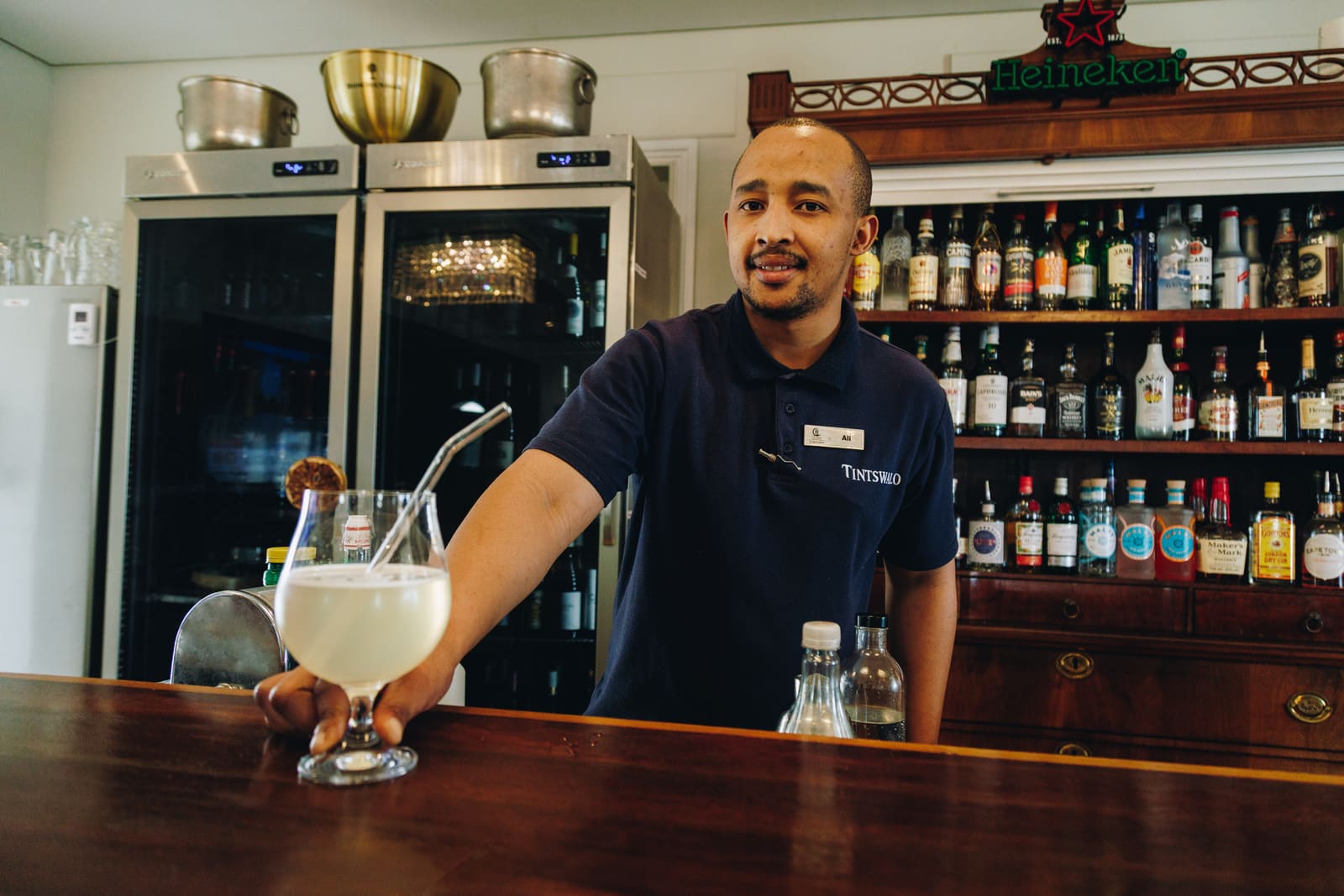 A smiling bartender in a navy polo shirt sliding a creamy cocktail with a straw across a wooden bar, with shelves of spirits behind him