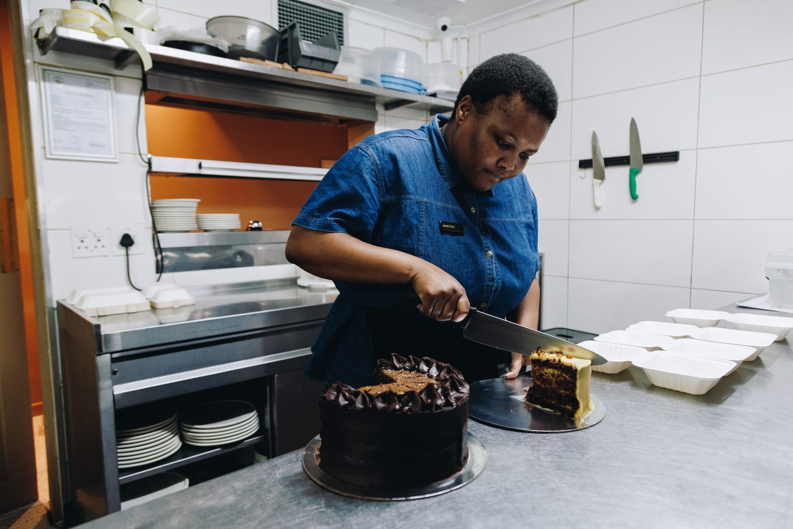 A hospitality worker in a denim shirt slicing a chocolate cake in a kitchen, takeaway boxes waiting beside her
