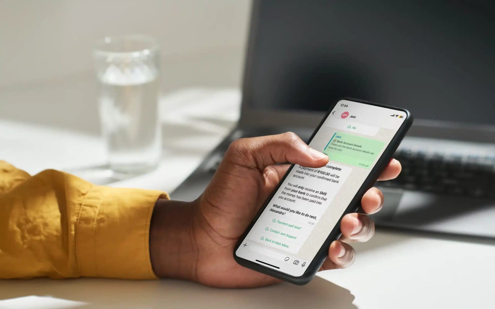 A hand holding a phone with a Jem WhatsApp conversation open, at a desk