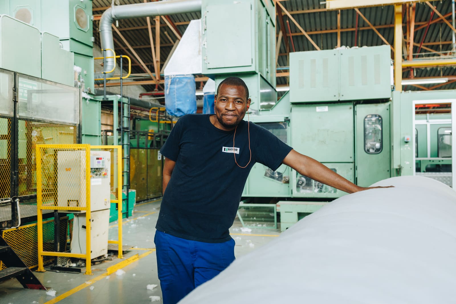 A factory worker in a navy t-shirt and blue trousers standing with one arm resting on a large roll of white material in front of green production machinery