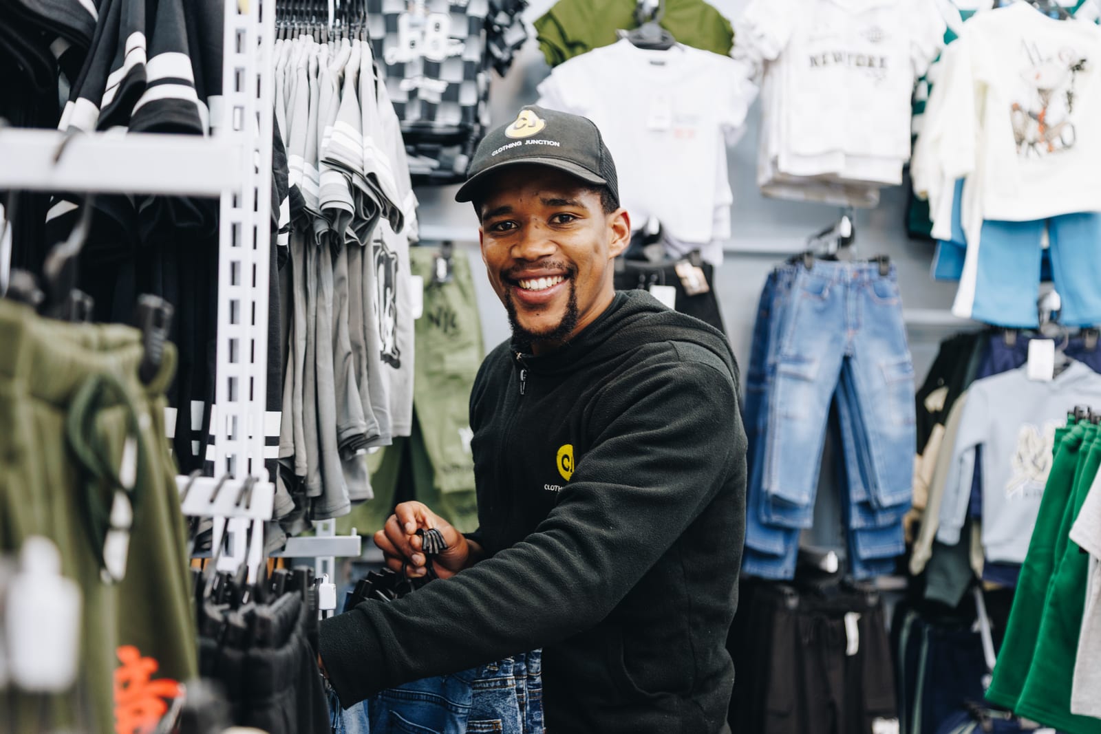 A shop assistant in a branded fleece and cap smiling beside rails of clothing