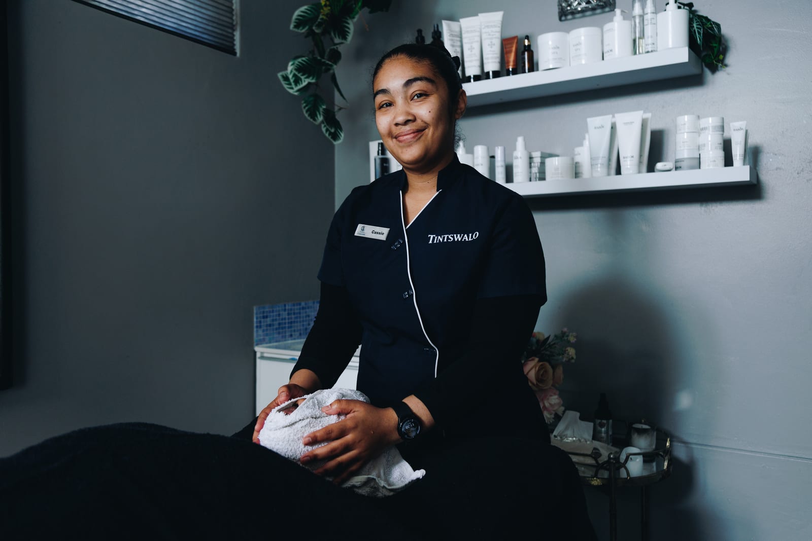 A spa therapist in a black Tintswalo tunic smiling beside a treatment bed, shelves of skincare products on the wall behind her