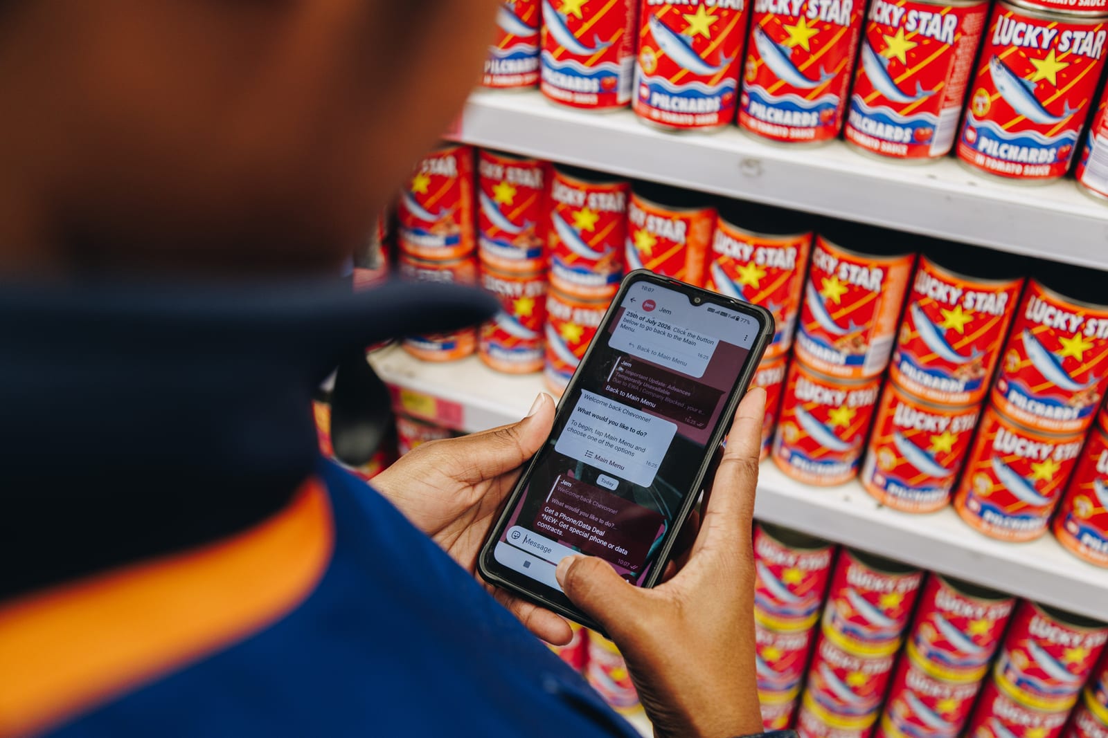 A supermarket worker reading a Jem WhatsApp menu on her phone in front of shelves of tinned fish