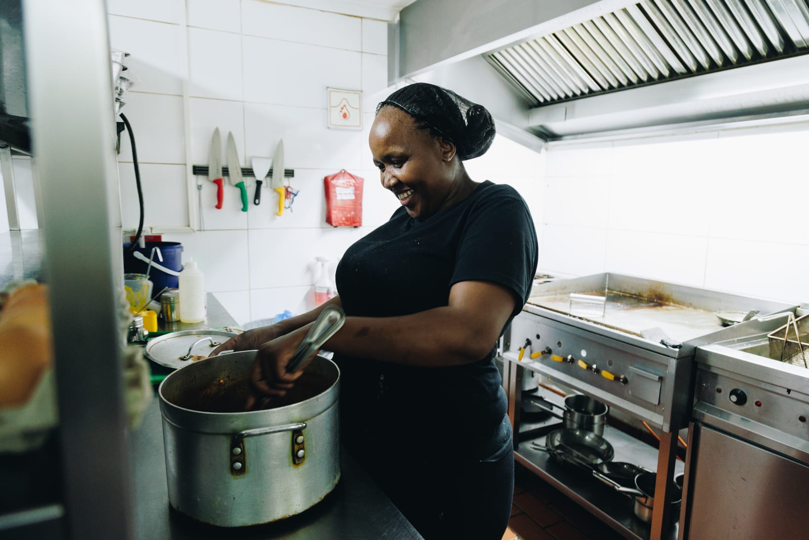 A kitchen worker in a hairnet smiling as she stirs a large pot on a stainless steel counter, with knives and a griddle station behind her