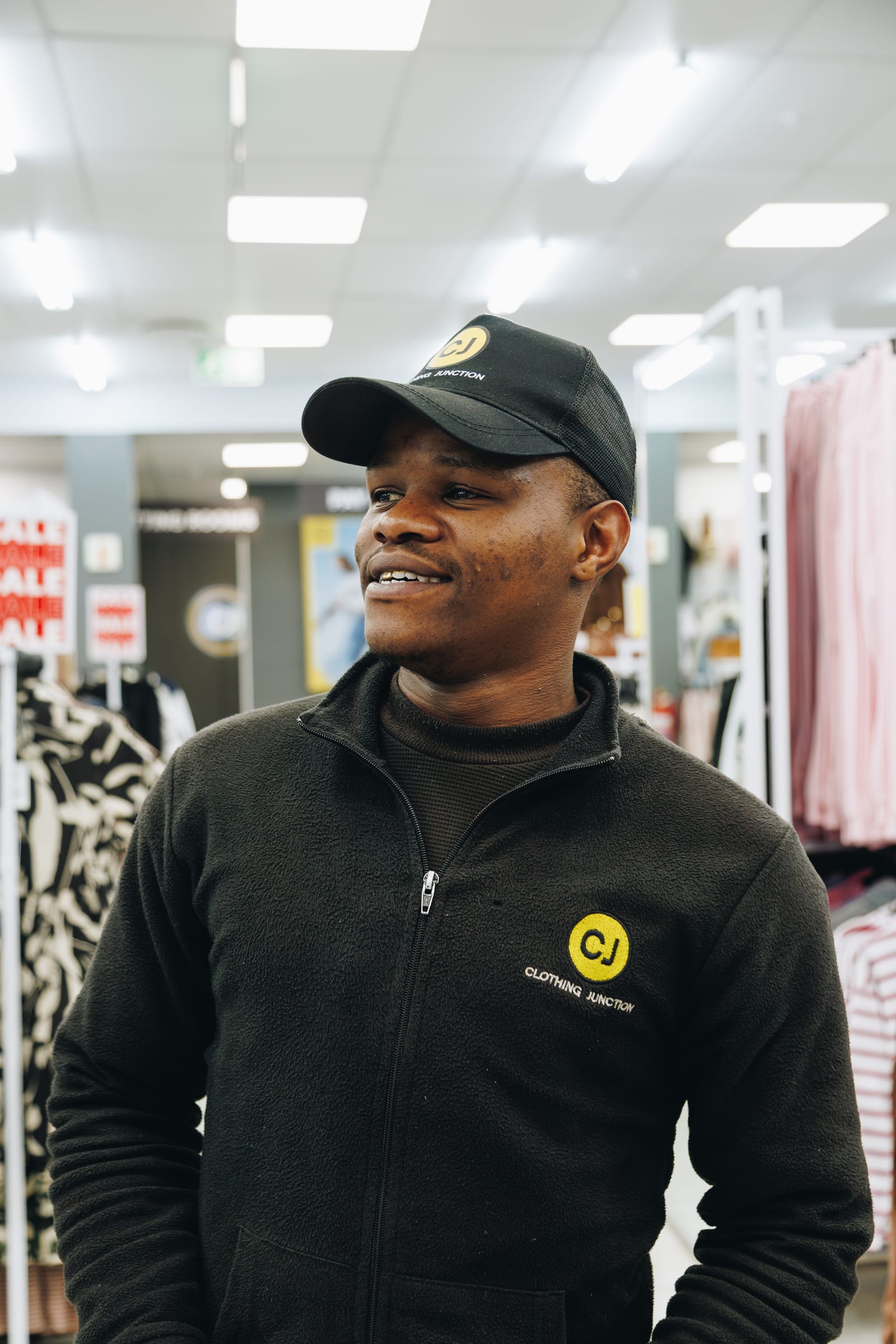 A shop assistant in a branded fleece and cap looking off to one side, clothing rails behind him