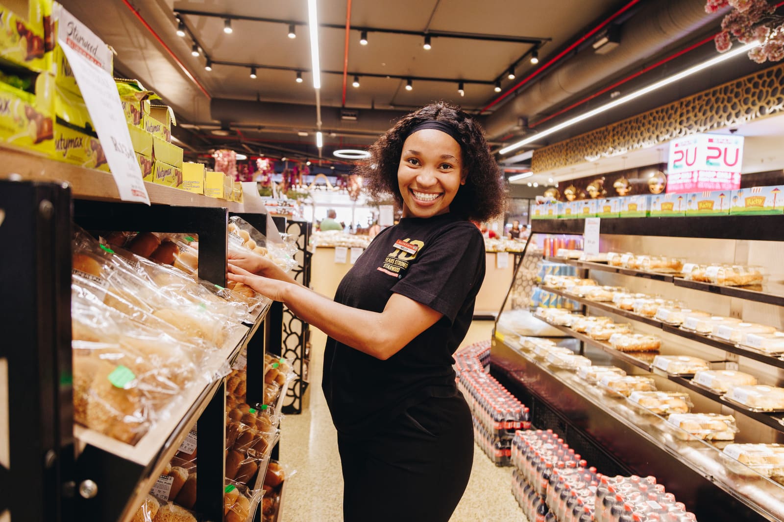 A smiling supermarket assistant in a black uniform stacking fresh bread on a shelf in the bakery aisle