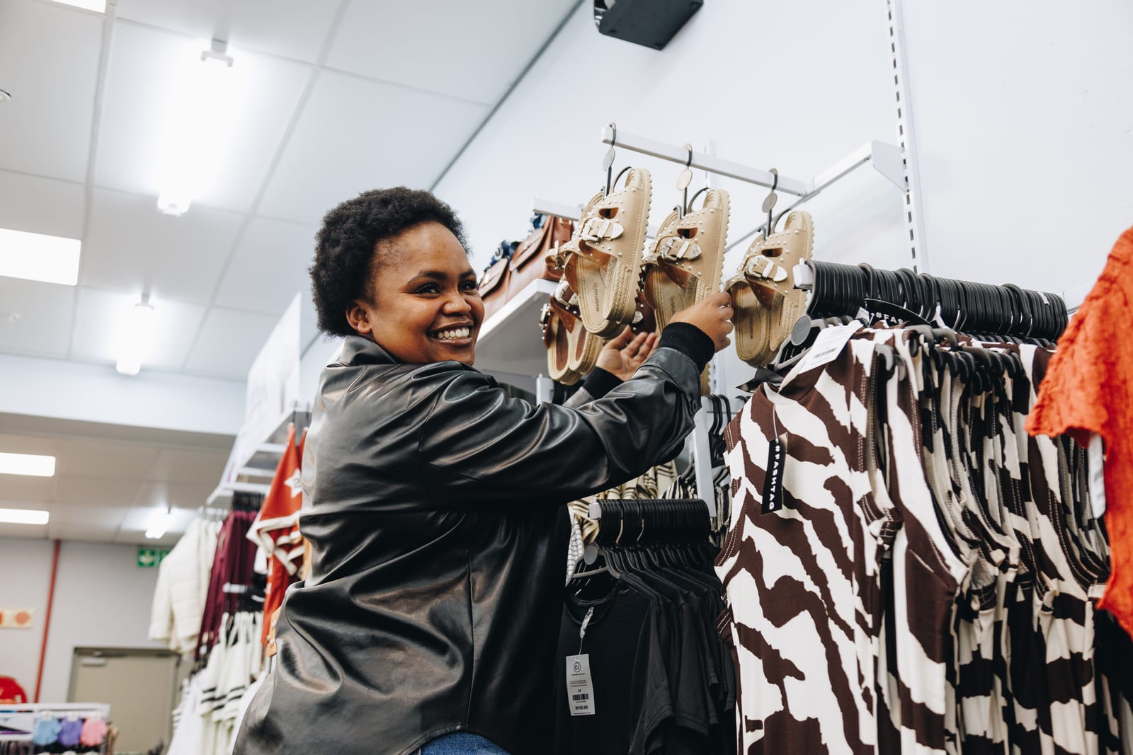 A shopper in a leather jacket smiling as she reaches for a pair of sandals on a shop display
