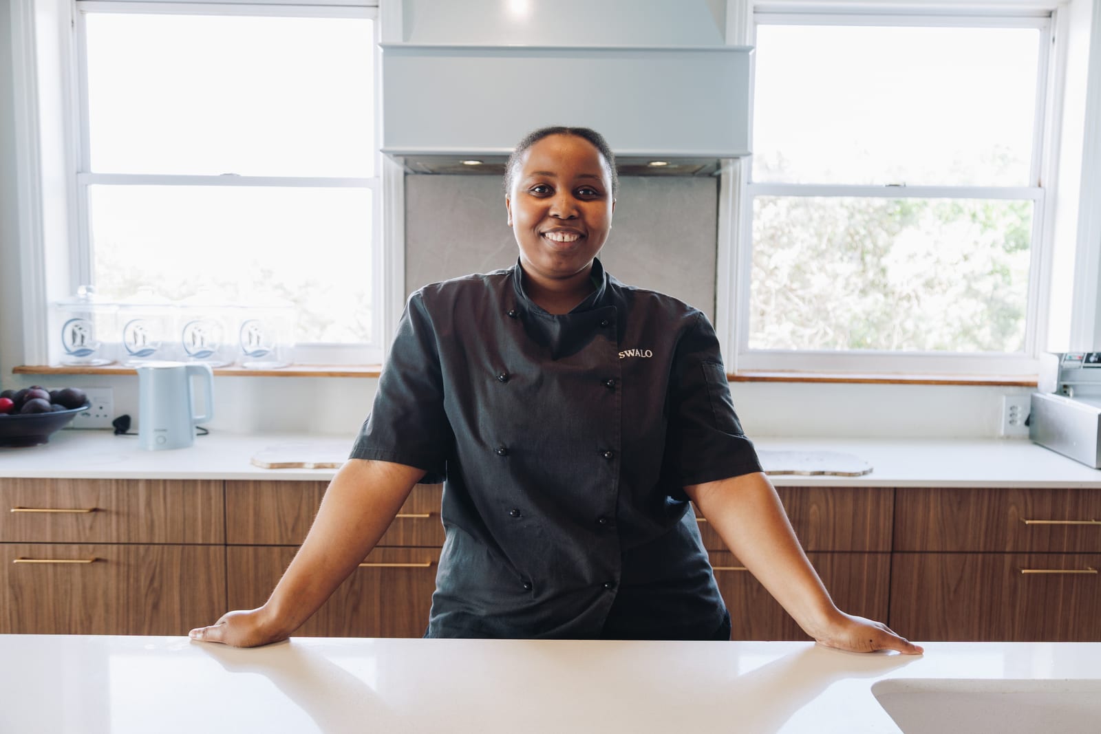 A chef in black kitchen whites standing behind a kitchen counter, hands on the worktop, smiling at the camera