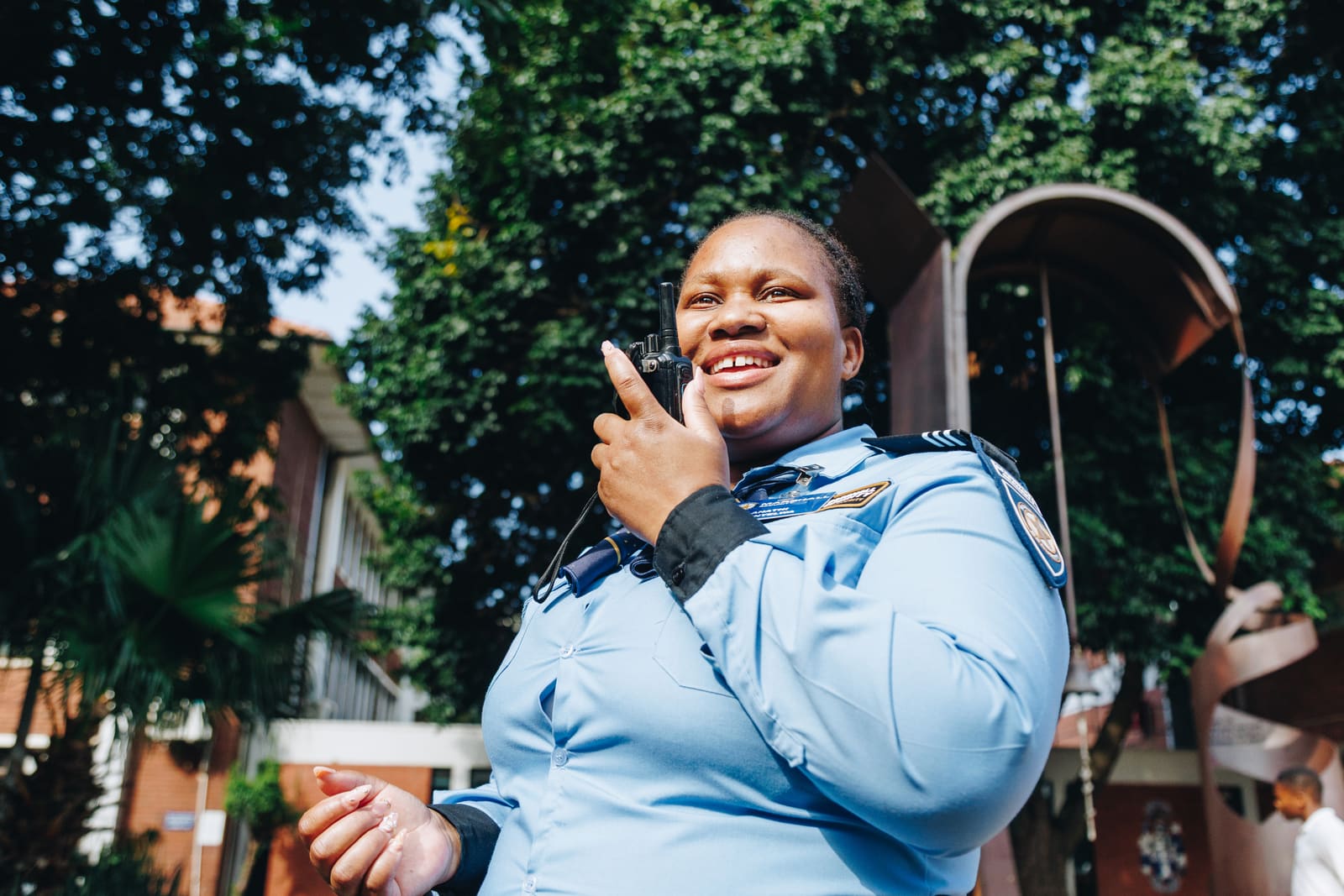 A smiling security officer in a light blue uniform speaking into a handheld radio outside a campus building
