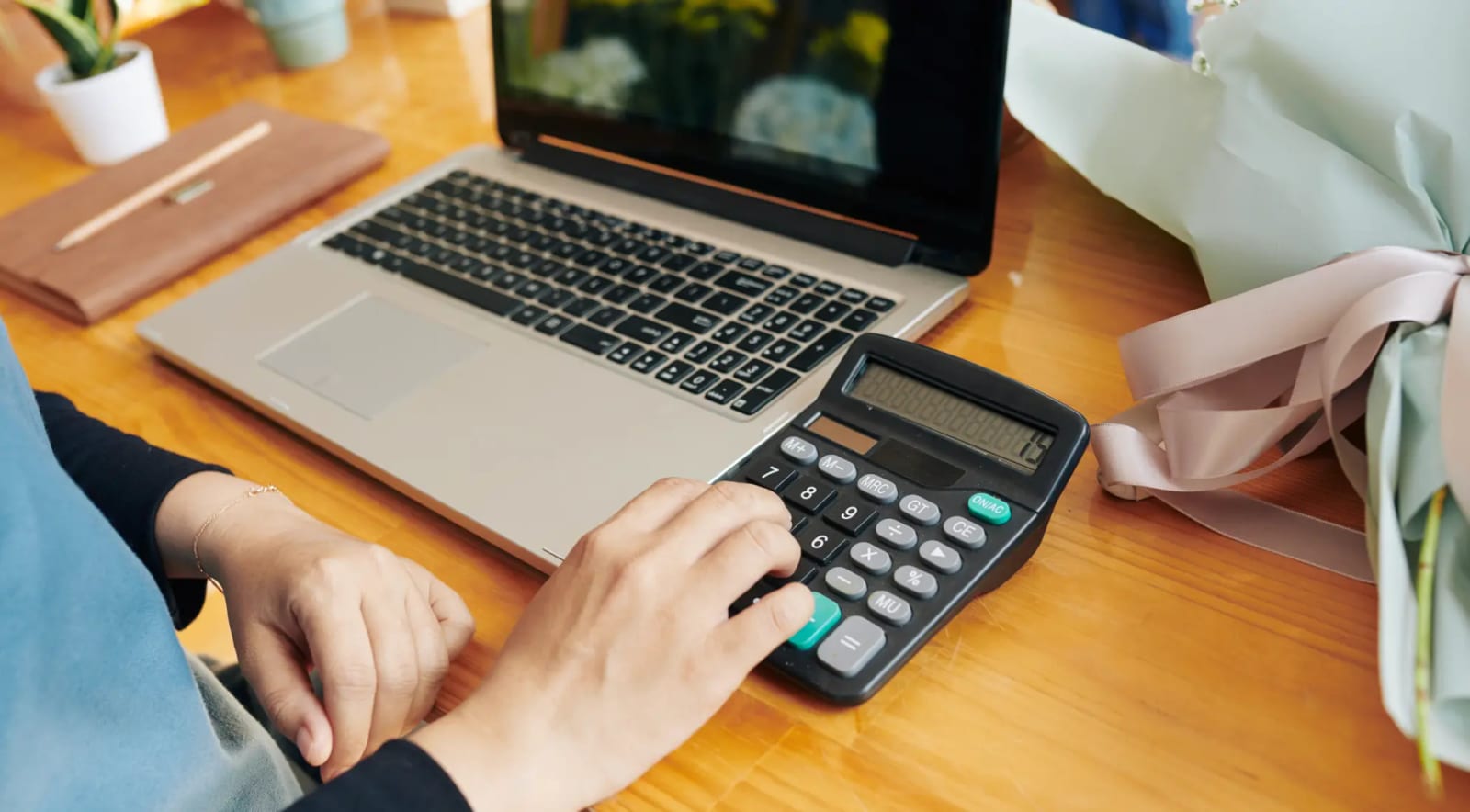 A hand working a desk calculator beside a laptop, with wrapped flowers on the table