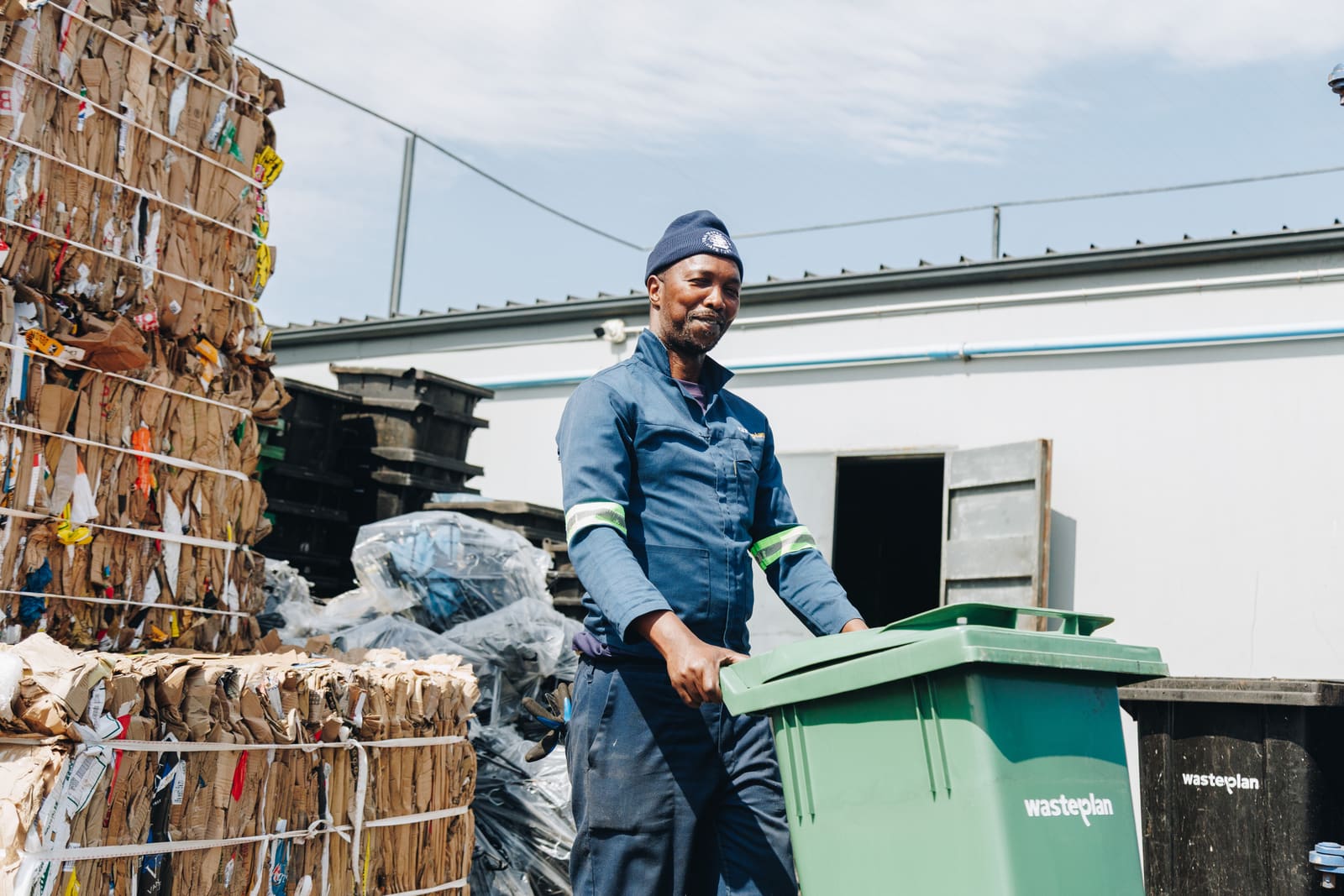 A smiling waste collector in a blue uniform and beanie wheeling a green wheelie bin past bales of baled cardboard at a recycling yard